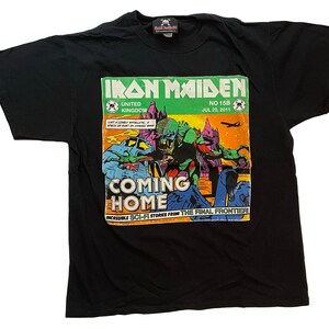 May include: Black t-shirt featuring a comic book-style graphic with the text "IRON MAIDEN" and "COMING HOME." The graphic includes a monster, a spaceship, and the date July 20, 2011. The shirt has a tag at the top.
