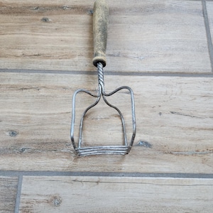 May include: A vintage metal potato masher with a wooden handle. The masher has a twisted wire design and a rectangular shape.