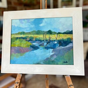 Könnte beinhalten: Originales Ölgemälde einer Küstenlandschaft, gerahmt in einem weißen Holzrahmen. Das Kunstwerk zeigt einen blauen Wasserweg mit Booten, grünes Marschland und einen blauen Himmel mit Wolken. Das Gemälde wird auf einer hölzernen Staffelei ausgestellt.