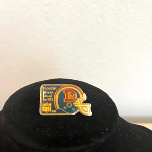 May include: A rectangular metal pin featuring a teal football helmet with an orange letter D logo. The pin includes the text Kodak Official Film of the NFL and the NFL logo in gold, blue, and white colours with a gold-toned metal border outline.