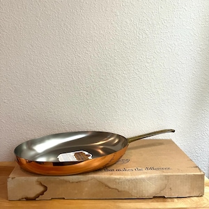 May include: A round copper frying pan with a silver-toned interior and a long brass handle. The pan rests on a rectangular cardboard box featuring the printed text, "...are that makes the difference." A small white product label is inside the pan.