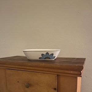 May include: A white ceramic serving bowl with a blue and green floral design. The bowl is sitting on a wooden surface.