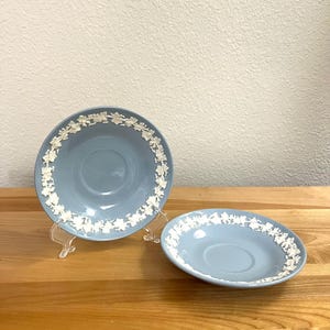 May include: Two light blue saucers with a white floral design around the rim. One saucer is on a clear plastic stand. The saucers are on a wooden surface.