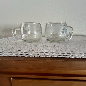 May include: Two clear glass mugs with a world map design. The mugs have a handle and are sitting on a white lace doily.