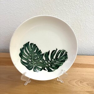 May include: A round white ceramic plate featuring a detailed graphic print of two dark green monstera leaves positioned at the bottom center. The plate is supported by a clear plastic stand, showcasing a clean, smooth surface and a simple design.