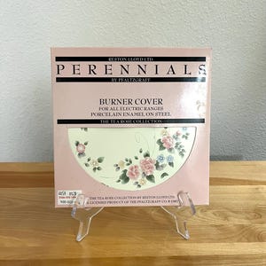 May include: A pink box displaying a burner cover for electric ranges. The cover features a floral design with pink, blue, and white flowers. Text on the box reads "PERENNIALS" and "THE TEA ROSE COLLECTION."