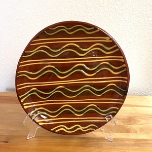 May include: A round ceramic plate with a glossy dark brown glaze featuring horizontal patterns of raised yellow wavy lines and straight yellow lines across the surface, displayed on a clear plastic stand for upright viewing.