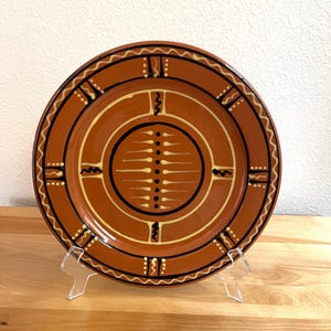 May include: A round terracotta-colored ceramic plate featuring a central circular design with a vertical row of black dots and radiating white lines. The plate is decorated with black concentric rings, wavy white lines, and geometric accents.