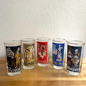 May include: A set of five clear glass tumblers with gold rims, each featuring a colorful graphic illustration of a city and its cultural icon. The glasses are labeled Rome, Bombay, Madrid, Athens, and The Hague, showcasing distinct regional designs.