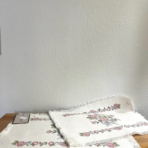 May include: Two rectangular off-white fabric placemats featuring a floral pattern with pink roses, green leaves, and blue accents. Each placemat has a fringed edge on all sides and a woven texture. One placemat includes a small rectangular paper tag.