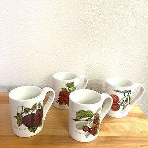 May include: Four white ceramic mugs with fruit illustrations. Each mug features a different fruit: plums, red currants, cherries, and peaches. The mugs have curved handles and are set on a wooden surface.