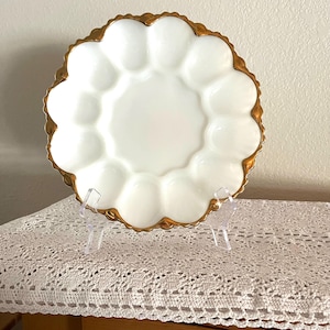 May include: A white, scalloped egg serving plate with a gold-colored trim. The plate has 12 egg-shaped indentations around a central circular area. The plate is displayed on a clear stand, and the background is a neutral color.