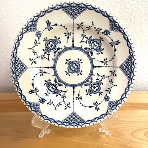 May include: A white ceramic plate with a blue floral design. The plate features a detailed pattern of flowers, leaves, and geometric shapes. The rim has a scalloped edge. The plate is displayed on a clear plastic stand.