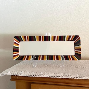 May include: Rectangular serving platter with a white center and a colorful striped border in shades of red, yellow, green, and black. The platter is displayed on a clear stand, showcasing its design.