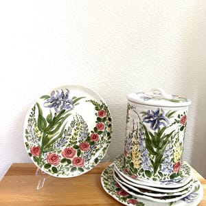 May include: A ceramic set featuring a round plate and a lidded canister, both decorated with hand-painted purple irises, yellow lupines, and pink roses on a white background. The canister sits atop a stack of matching plates.