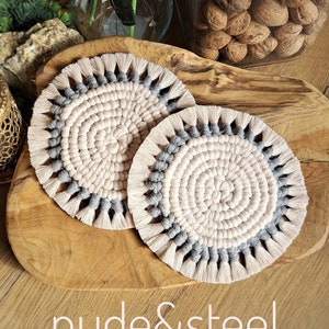 以下が含まれることがあります： 薄いピンクの中心と灰色の外側の輪を持つ、2つの丸い編み物のコースター。コースターは縁にフリンジが付いています。コースターは木製の表面にあります。