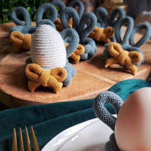 May include: A collection of handmade Easter egg holders. Each holder is crafted from knitted yarn in shades of gray and mustard yellow, featuring a bow detail. The holders are designed to cradle eggs, with a bunny ear design.