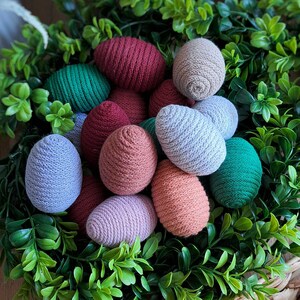 May include: A collection of colourful crocheted Easter eggs in shades of pink, red, green, and blue, nestled in a bed of green foliage.