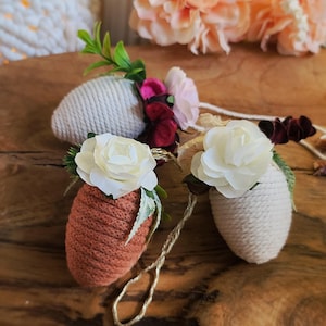 Può includere: Tre uova di Pasqua decorative, ciascuna avvolta in filo bianco e sormontata da un fiore bianco. Un uovo è marrone e gli altri due sono bianchi. Le uova sono appese a una corda.