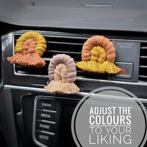 Può includere: Tre profumatori per auto in macramè colorati appesi alla bocchetta di ventilazione di un'auto. I profumatori sono in tonalità di arancione, giallo, marrone, rosa e beige. Il testo "ADJUST THE COLOURS TO YOUR LIKING" è visibile nell'immagine.
