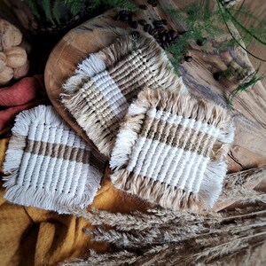 May include: Three handmade macrame coasters with a white and brown striped pattern. The coasters are made with natural fibers and have a fringed edge.