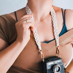 Puede incluir: Una persona lleva un top marrón sin mangas con una correa de cámara de crochet blanco. La correa está unida a una cámara vintage negra con el texto "LOMO 135 B" en la parte delantera.