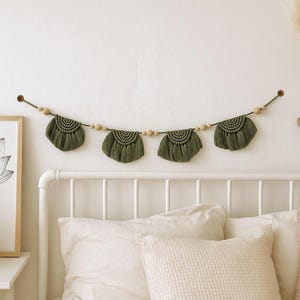 May include: A decorative wall hanging featuring four olive green macrame fan shapes, strung together with wooden beads. The garland is suspended above a white bed frame with white bedding. A framed botanical print and a vase of dried flowers are also visible.