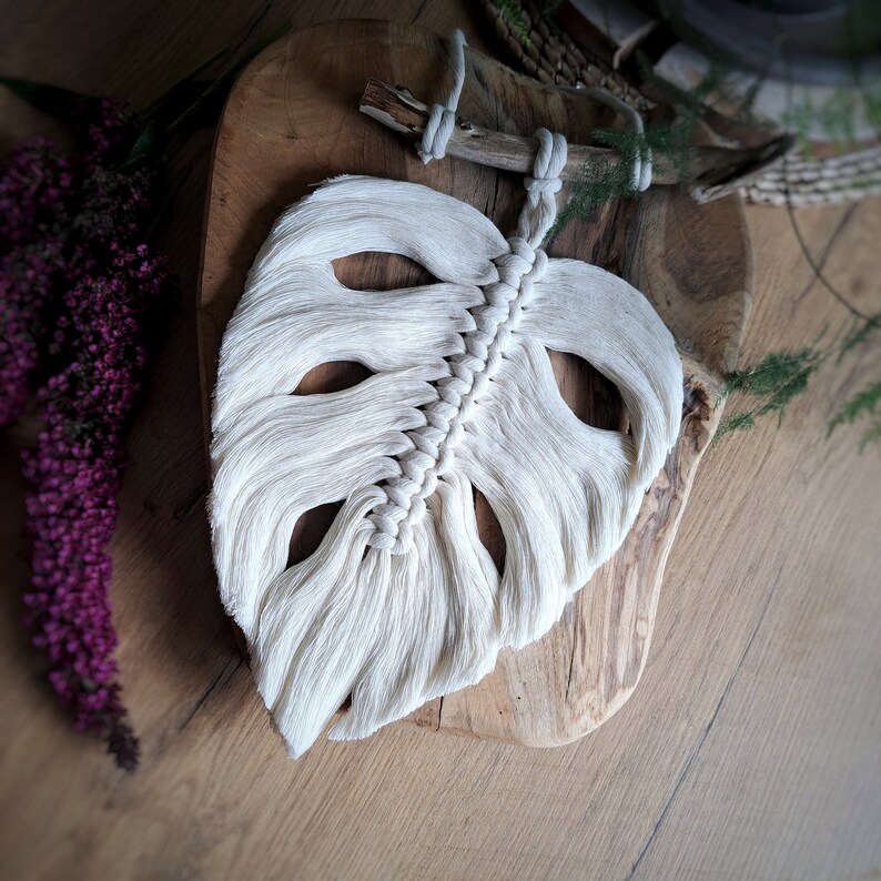 May include: A white macrame wall hanging in the shape of a monstera leaf. The leaf is hanging on a wooden board.