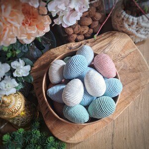 Op de afbeelding: Een kom gevuld met kleurrijke, met garen omwikkelde paaseieren. De eieren zijn in tinten blauw, wit en roze. De kom is van hout en heeft een natuurlijke afwerking.