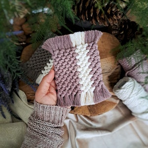 Puede incluir: Un posavasos de macramé hecho a mano con un patrón geométrico en tonos rosa y blanco. El posavasos tiene un borde de flecos y está hecho de fibras naturales.