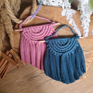 May include: Two macrame wall hangings, one dusty rose and one teal, are displayed on a wooden surface. Each features a half-circle design with a fringe of knotted threads. A wooden dowel supports the top, and a loop is attached for hanging.
