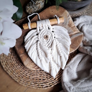 Puede incluir: Un tapiz de macramé blanco con un diseño circular en el centro y una barra de madera en la parte superior. El tapiz está hecho de cordón de algodón y tiene un borde con flecos.