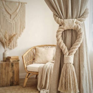 Può includere: Tende in lino beige tenute da un fermatenda in corda spessa color crema con nappa. Davanti alle tende c'è una sedia in vimini con un cuscino e una coperta color crema. Sono visibili anche un arazzo in macramè e un vaso.