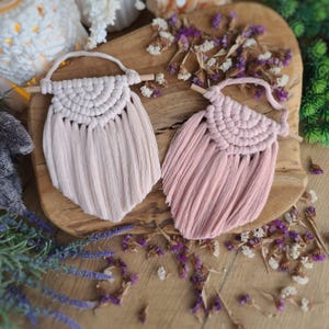 May include: Two pink macrame wall hangings with wooden dowels and fringe. The hangings are in a light pink and a darker pink.