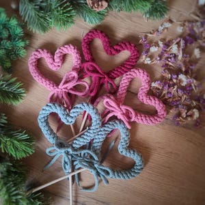 Puede incluir: Decoraciones artesanales en forma de corazón, en tonos rosa y azul, cada una con un lazo. Los corazones están hechos de un material grueso y tejido, y están unidos a palitos de madera. El fondo incluye vegetación y flores secas.