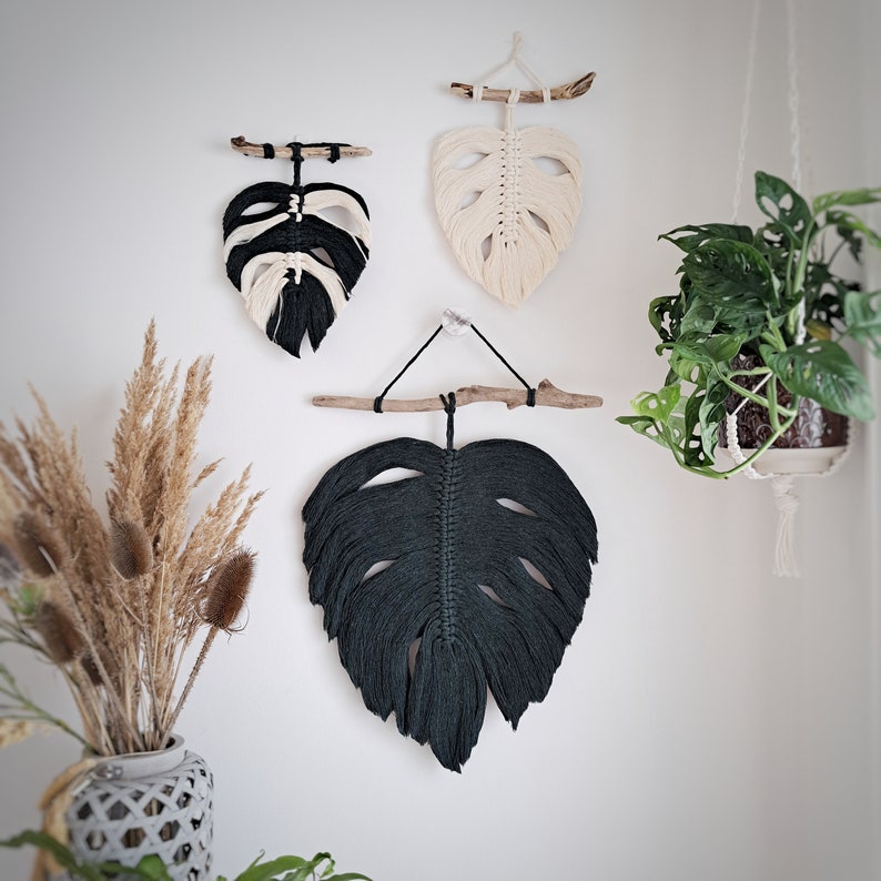 May include: Three macrame wall hangings in the shape of monstera leaves. One is white, one is black, and one is dark green.