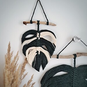 May include: Two macrame wall hangings in the shape of monstera leaves. The leaves are made with dark green and cream colored yarn and are attached to driftwood.