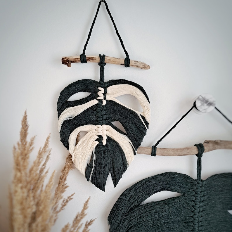 May include: Two macrame wall hangings in the shape of monstera leaves. The leaves are made with dark green and cream colored yarn and are attached to driftwood.