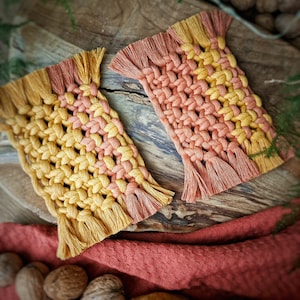 Puede incluir: Dos posavasos de macramé hechos a mano en un esquema de colores amarillo y naranja. Los posavasos tienen un borde de flecos y están hechos con un patrón de nudos tejidos.