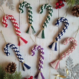 Könnte beinhalten: Eine Sammlung handgefertigter Weihnachtsornamente in Form von Zuckerstangen. Die Ornamente bestehen aus geflochtenem Garn in verschiedenen Farben, darunter Rot, Grün, Blau, Lila und Rosa, mit weißen Akzenten. Jede Stange hat eine Quaste und einen goldenen Faden zum Aufhängen.