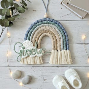 Op de afbeelding: Handgemaakte macramé regenboog wanddecoratie in blauwe, groene en beige tinten, met de naam "Gisele" in sierlijke letters. De regenboog heeft franjes aan de onderkant. Decoratie voor de kinderkamer.