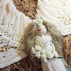 May include: A white yarn angel doll with gold yarn hair, a white crocheted dress, and a white flower crown. The doll is holding a bouquet of white flowers and is lying on a bed of brown shredded paper.