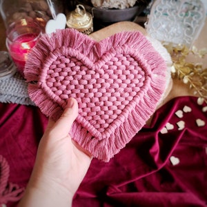May include: A handmade, pink macrame heart-shaped coaster with a fringed edge. The coaster is held in a hand, showcasing the intricate woven design. The coaster is a decorative item, perfect for Valentine's Day or home decor.