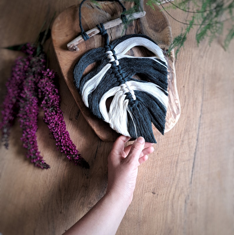 May include: A black and white macrame wall hanging in the shape of a monstera leaf. The leaf is hanging on a wooden board.