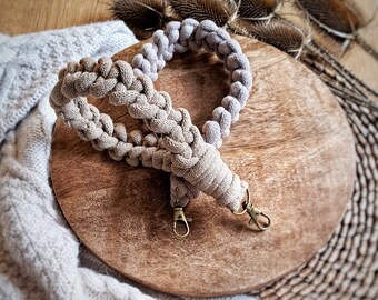 May include: A beige crocheted camera strap with two gold metal clasps. The strap is lying on a wooden surface.