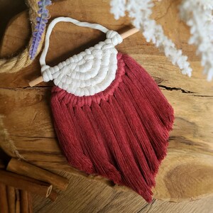 Puede incluir: Un colgante de pared de macramé hecho a mano con un diseño de semicírculo. La sección superior es blanca, con un patrón tejido, y la inferior es de color rojo intenso con flecos. Está suspendido de una clavija de madera con un cordón blanco.