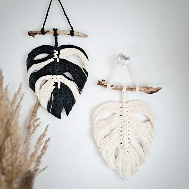May include: Two macrame wall hangings in the shape of leaves. One is black and white, the other is white. Both are hung from driftwood.