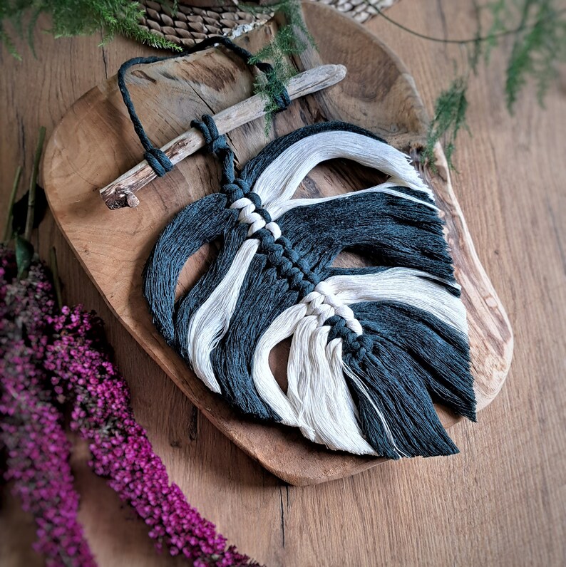 May include: A black and white macrame wall hanging in the shape of a leaf. The leaf is made with yarn and has a wooden dowel at the top.
