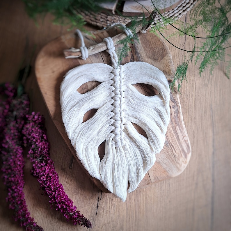 May include: White macrame wall hanging in the shape of a monstera leaf. The leaf is hanging on a wooden board.