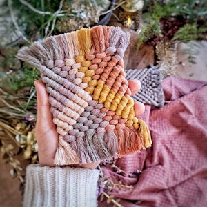 Può includere: Un piccolo arazzo da parete tessuto a mano con un motivo geometrico in tonalità di grigio, rosa, arancio e giallo. L'arazzo ha un bordo a frangia ed è realizzato con una tecnica di macramè.